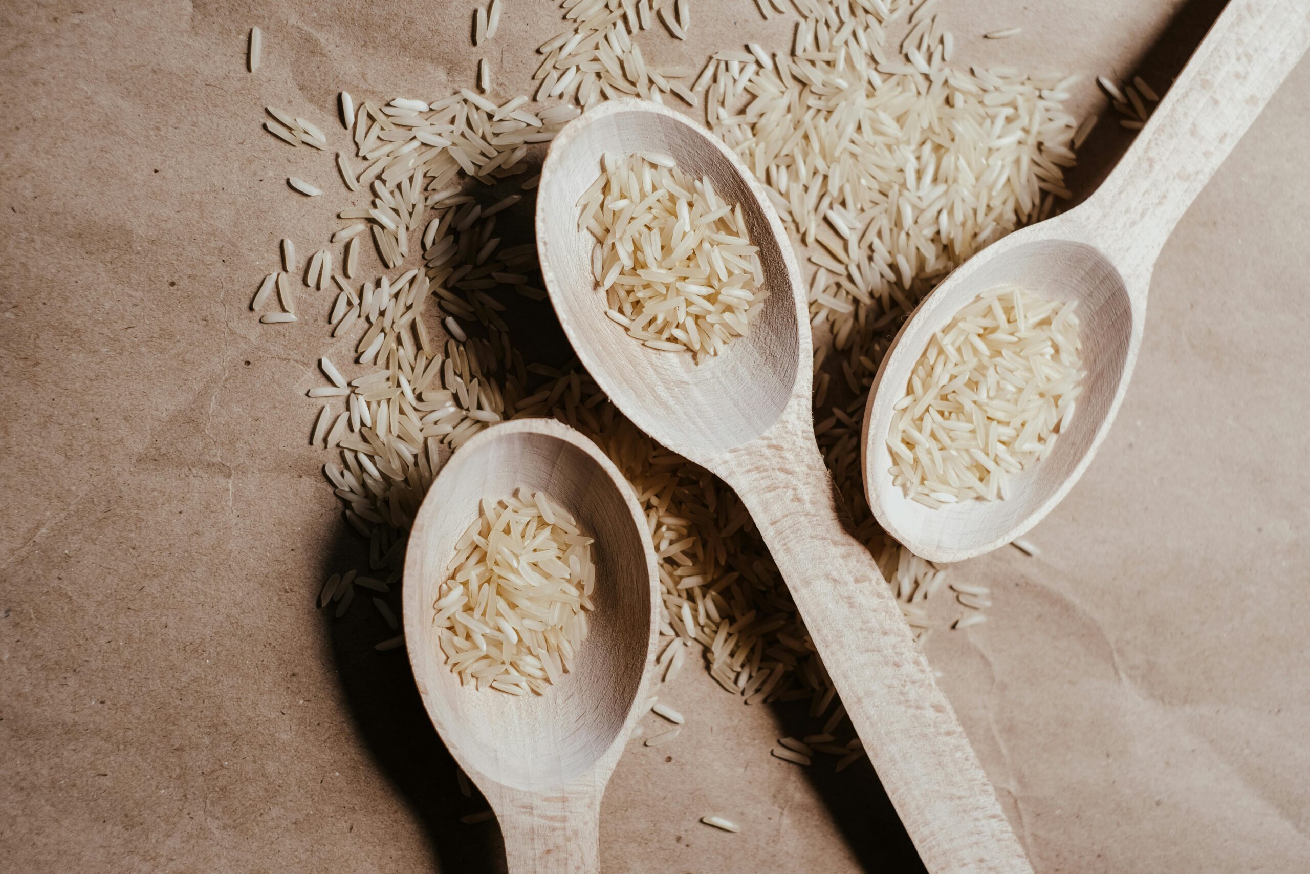 Three wooden spoons filled with uncooked rice on a textured brown paper surface.